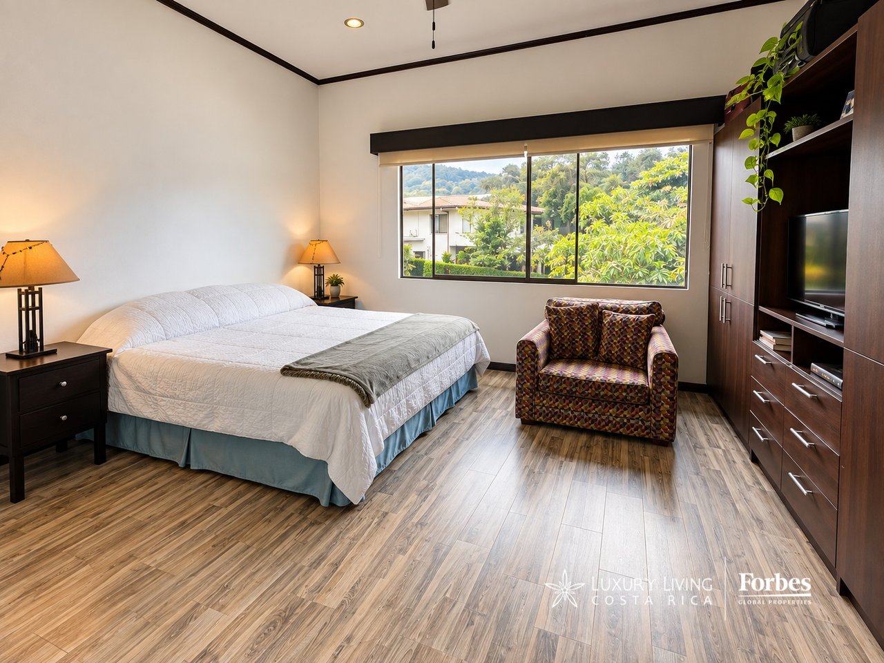 Furnished bedroom with a large bed, patterned armchair, dark built-in cabinetry, and a window overlooking greenery.