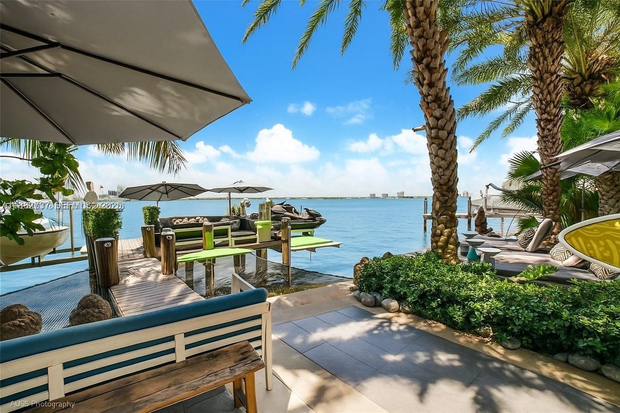 Waterfront patio with palm trees, shaded lounge seating, and a dock overlooking calm blue water.