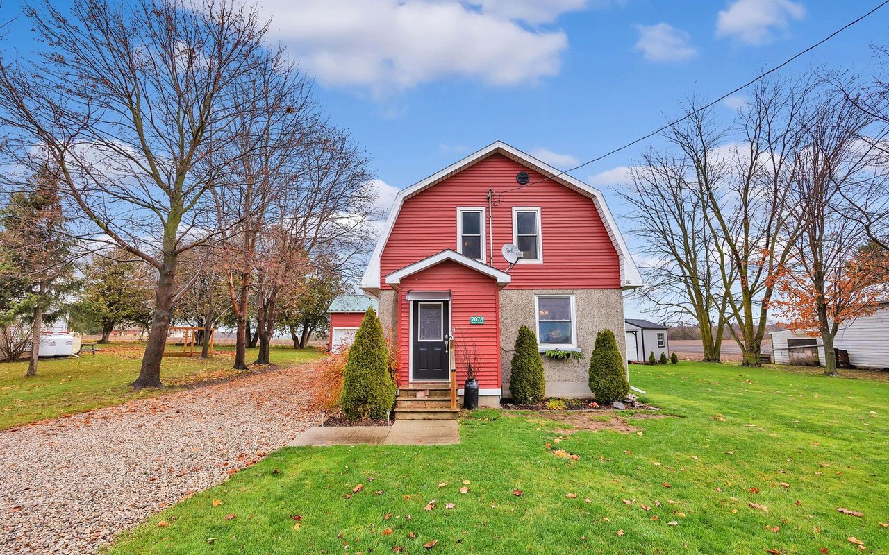Rural two-story house with red gambrel-style upper siding, stone lower level, and a gravel driveway.