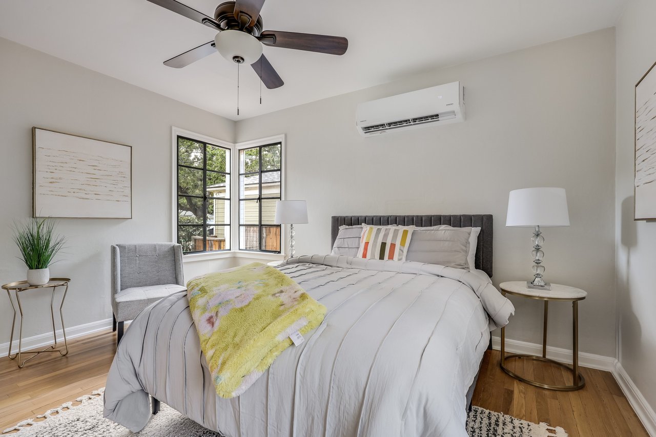 Staged bedroom with white bedding, a yellow floral throw, corner windows, and a ceiling fan.