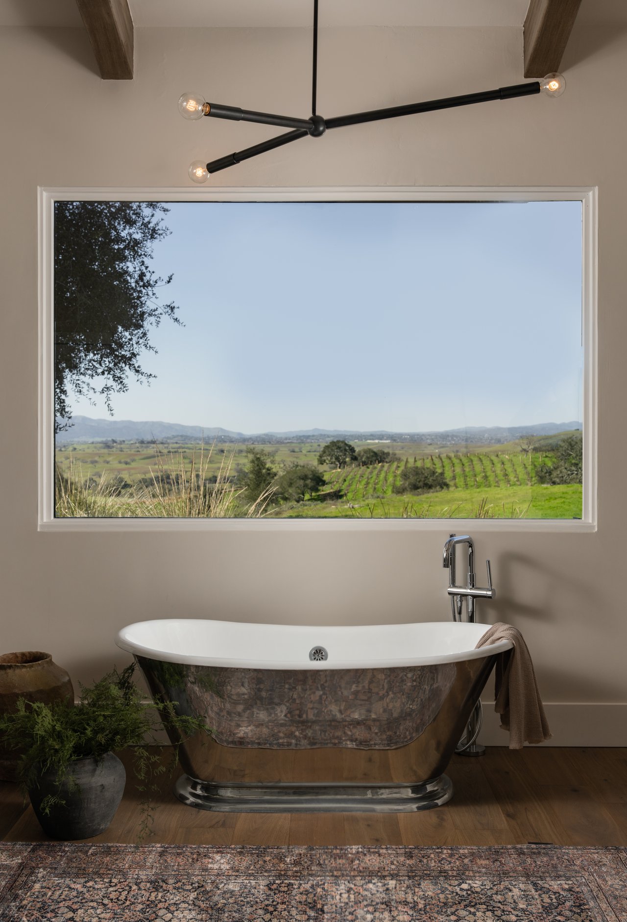 Freestanding bathtub beneath a picture window overlooking green vineyards and distant hills.