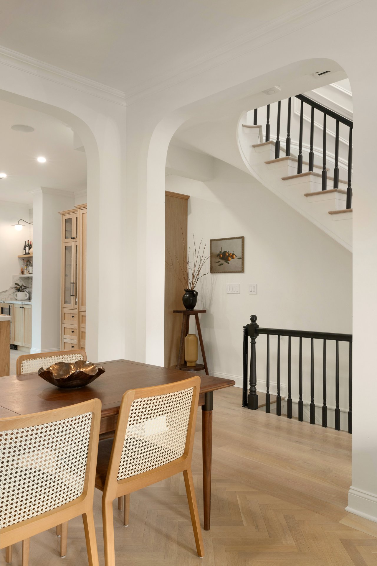 Dining area featuring a wooden table with cane-backed chairs and herringbone oak flooring, framed by white arched doorways leading to a classic staircase with black railings.