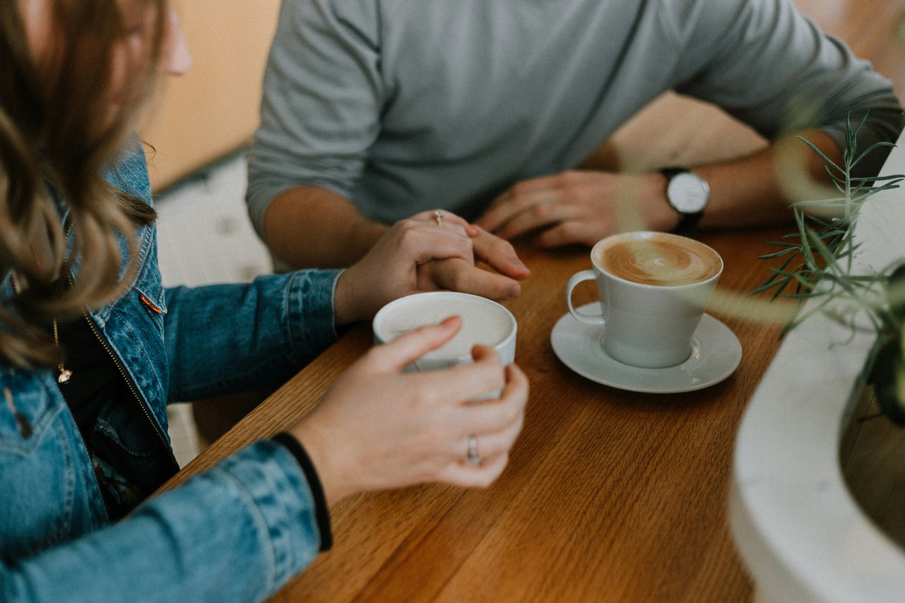 Two people hold hands over a café table beside cups of coffee.