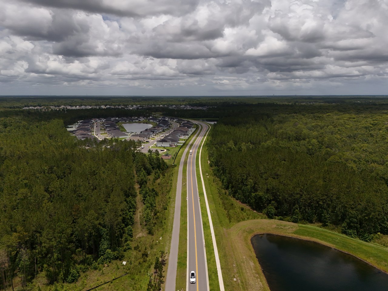 Aerial view of a neighborhood bordered by woodland, with a long road and pond in the foreground.