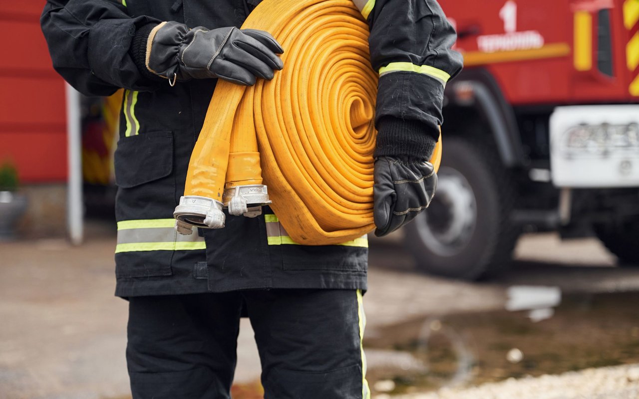 Firefighter in reflective gear holding a coiled yellow hose in front of a fire truck