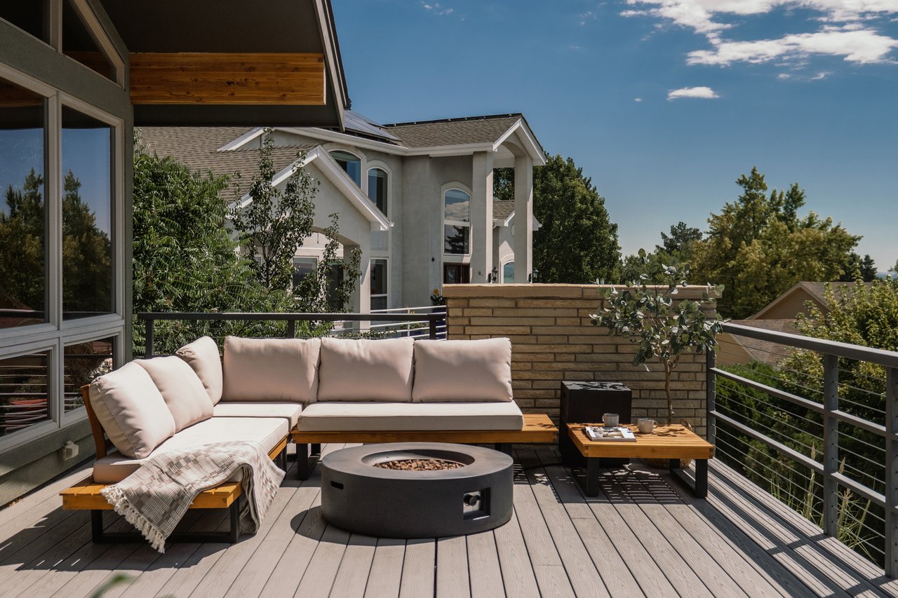 Outdoor deck with a sectional sofa and round fire pit, overlooking nearby homes and trees.