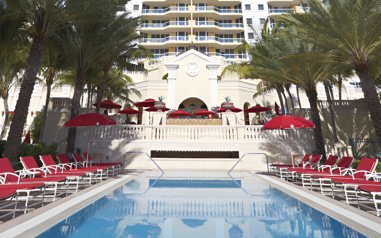 Resort pool lined with red lounge chairs and umbrellas beneath palm trees.
