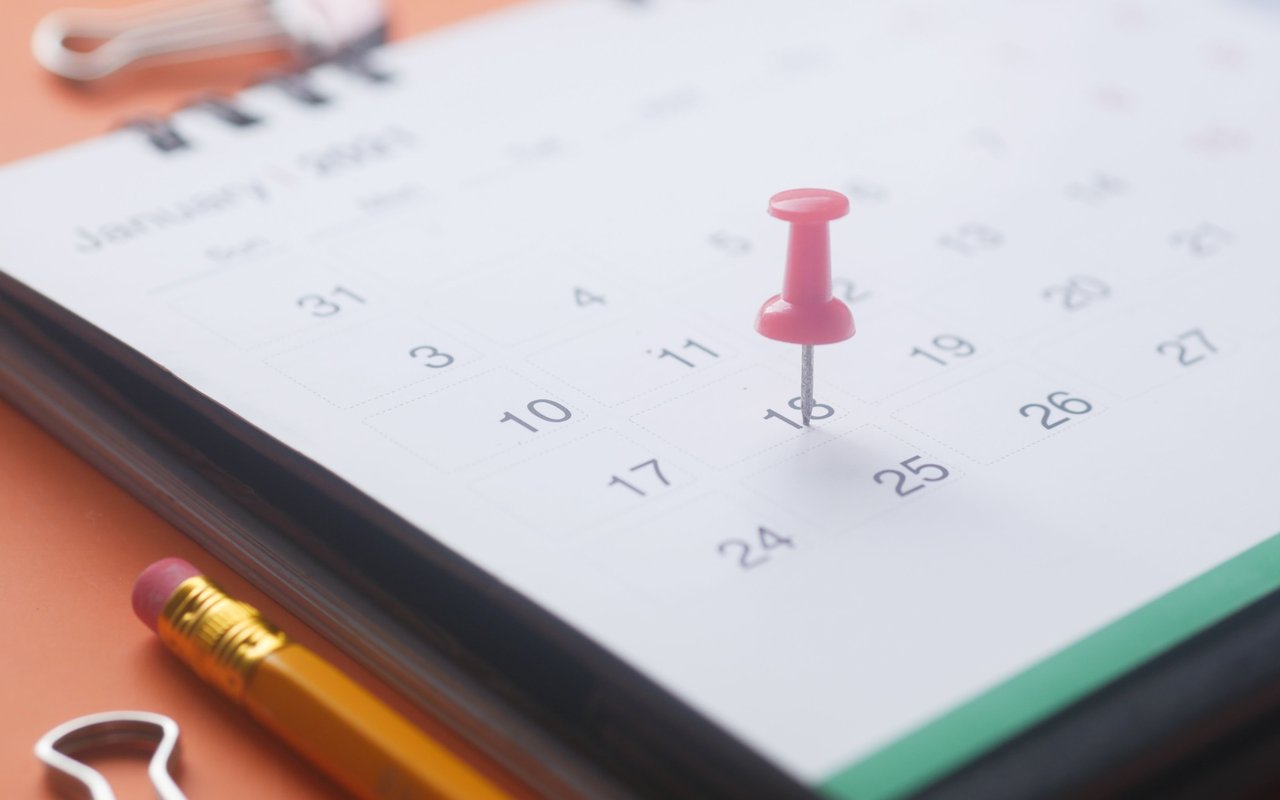 Pink pushpin marking a date on a calendar beside a yellow pencil and paper clips.