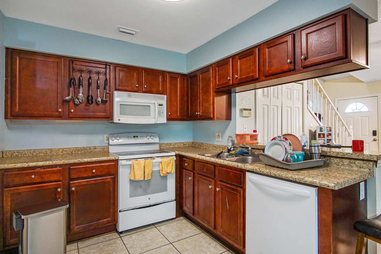 Kitchen at 2506 Nugget Lane in Tallahassee, Florida, featuring granite countertops, dark wood cabinets, and tile flooring. Appliances include a white electric range with overhead microwave, dishwasher, and stainless steel sink. A breakfast bar overlooks the living area with a view of the front entry and staircase.
