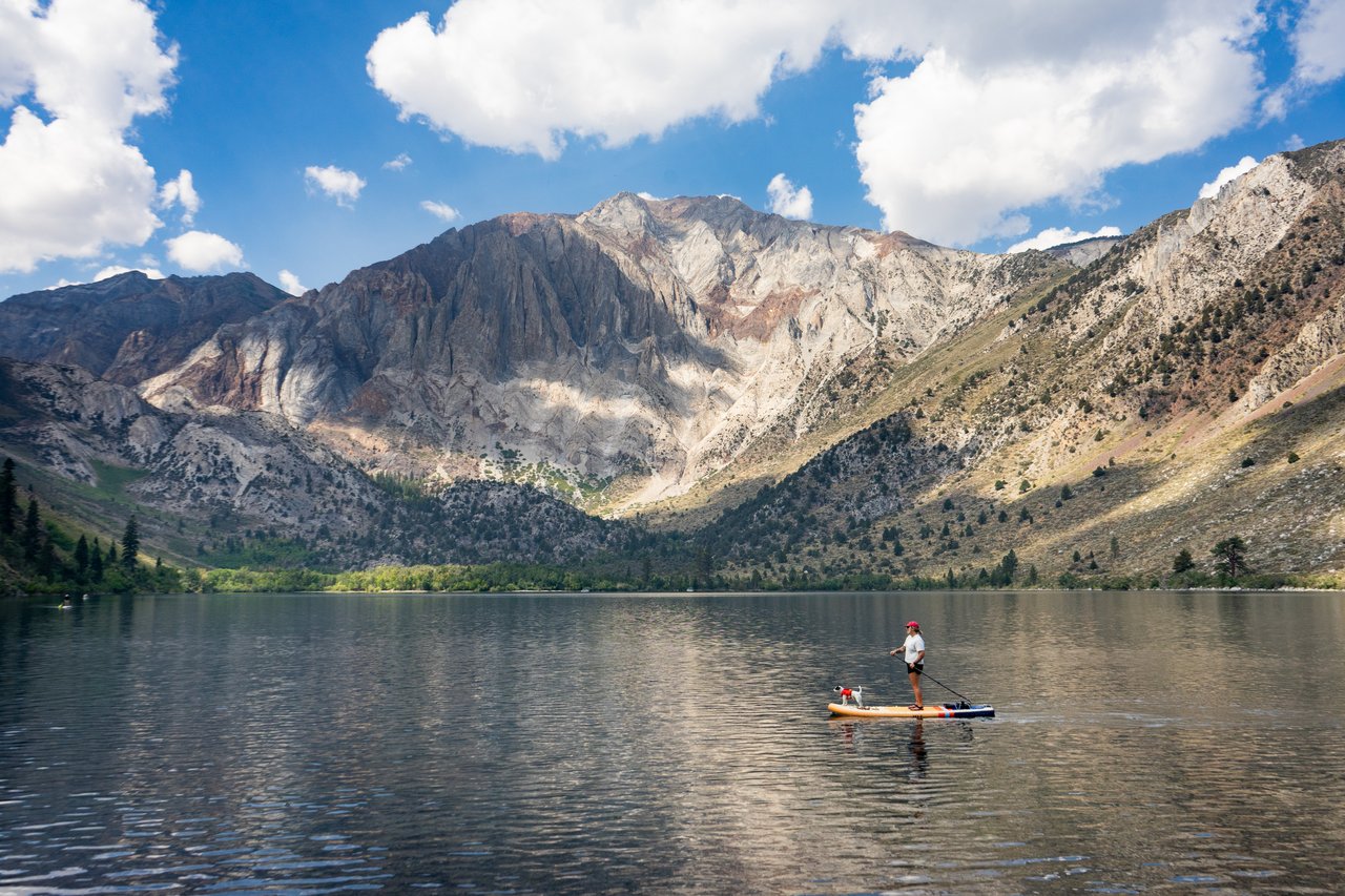 A paddleboarder crosses a mountain lake beneath rugged granite peaks and a cloud-filled sky.