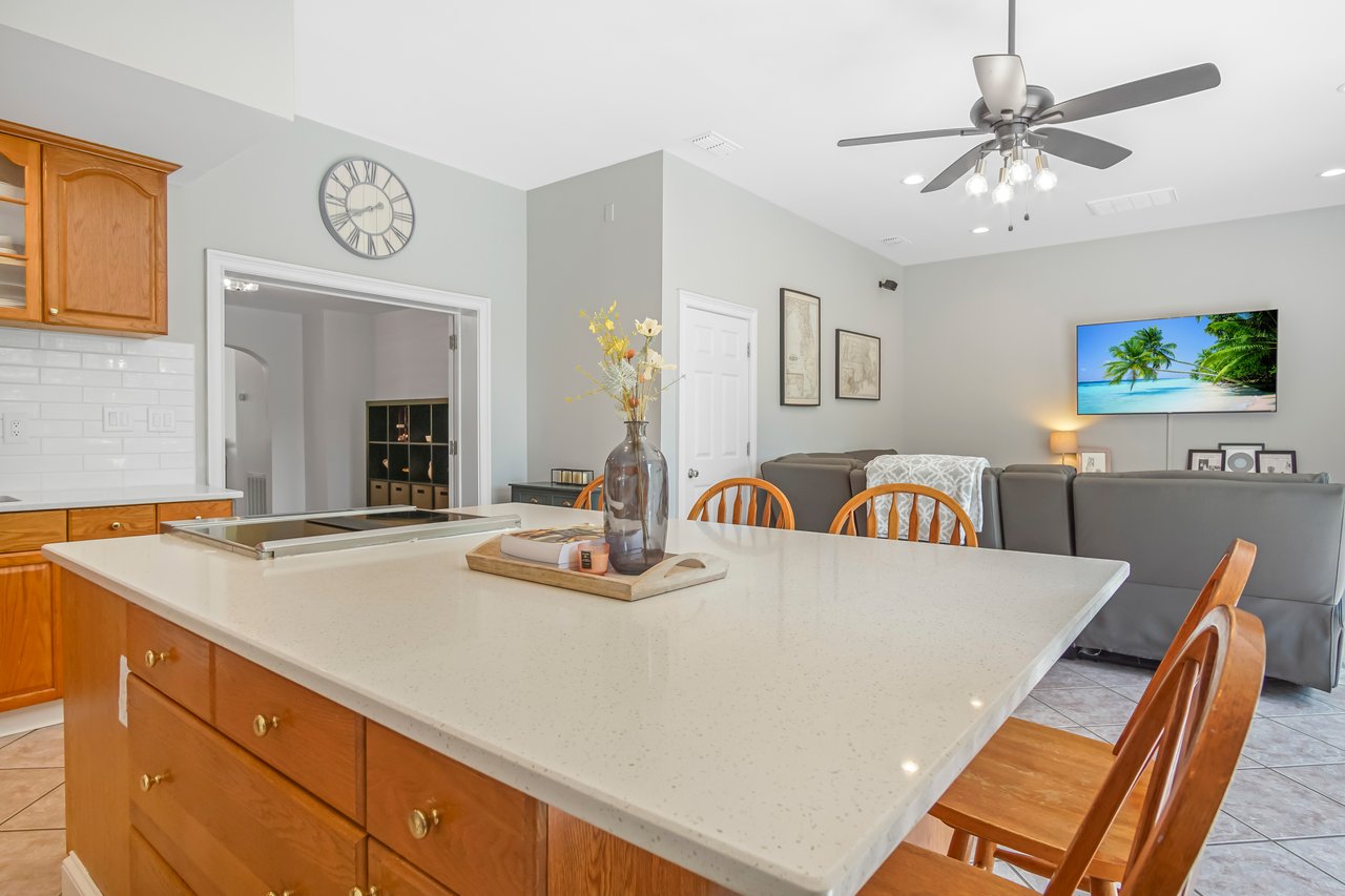 A modern kitchen with a large island, wooden cabinets, and a ceiling fan. A cozy living area features a sofa, tropical wall art, and a welcoming ambiance.