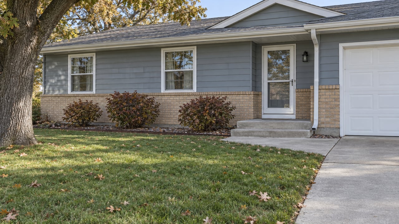 Blue-gray single-story home with buff brick, a white storm door, concrete front steps and a broad autumn lawn.