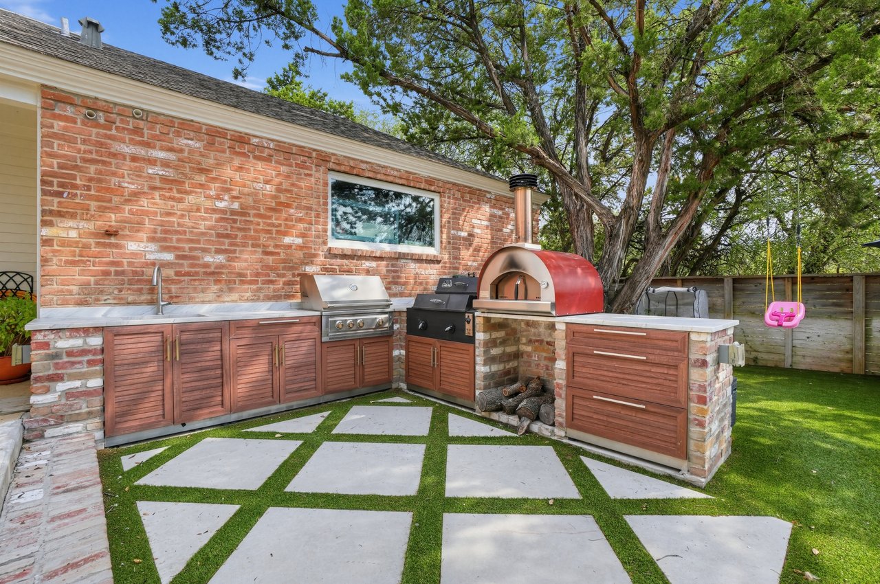 Brick outdoor kitchen with built-in grill, pizza oven, and stepping-stone patio beneath mature trees.