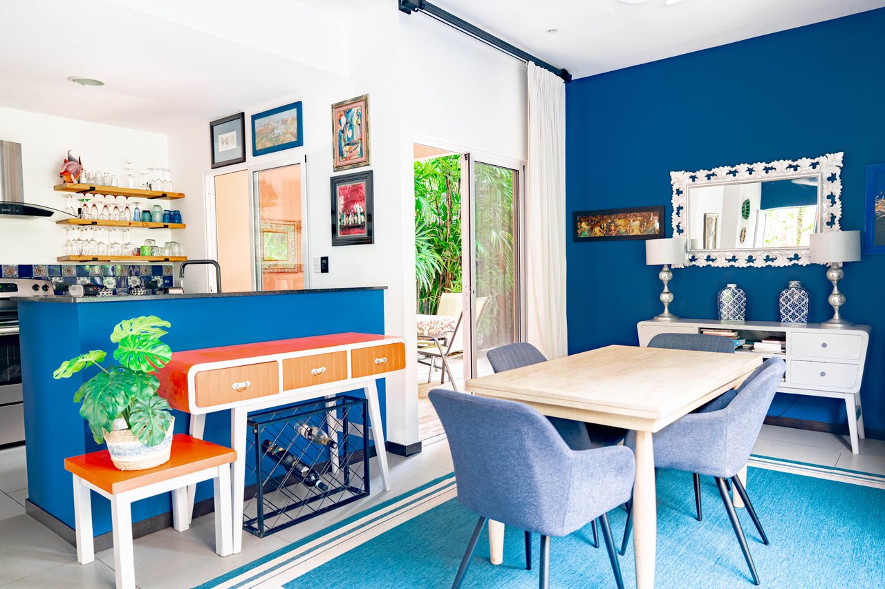 Dining area with blue chairs and a wooden table beside a vivid blue kitchen island and tropical greenery.