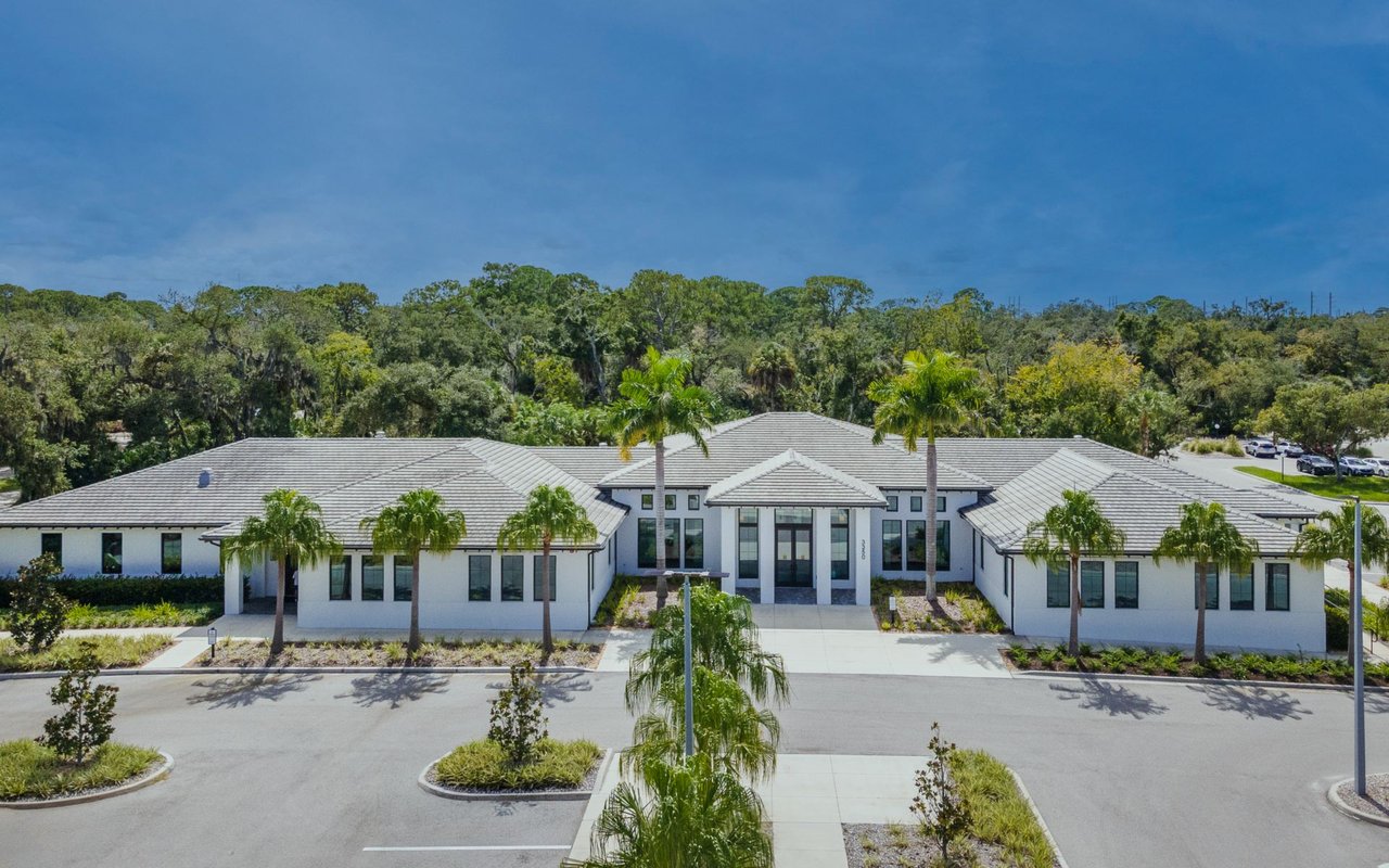 Low-rise white building with gray tiled roofs, palm trees, and a landscaped parking area.