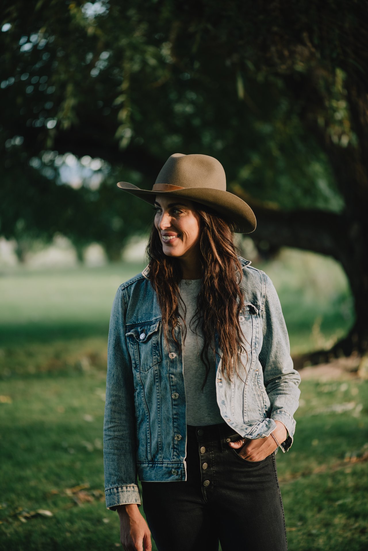 Smiling woman in a tan hat and denim jacket standing among trees.