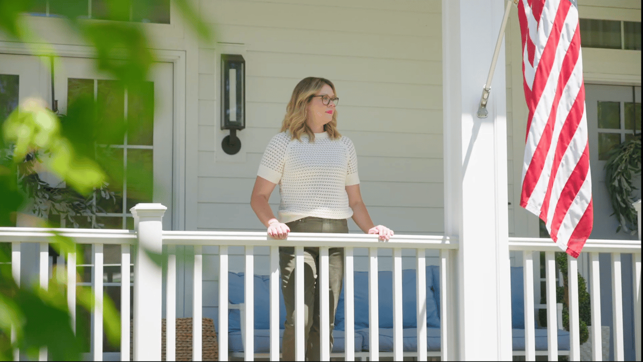 A Madison homeowner standing on her white front porch on a sunny morning, looking out over her neighborhood, representing the long-term homeowners building equity by staying in their Madison homes.