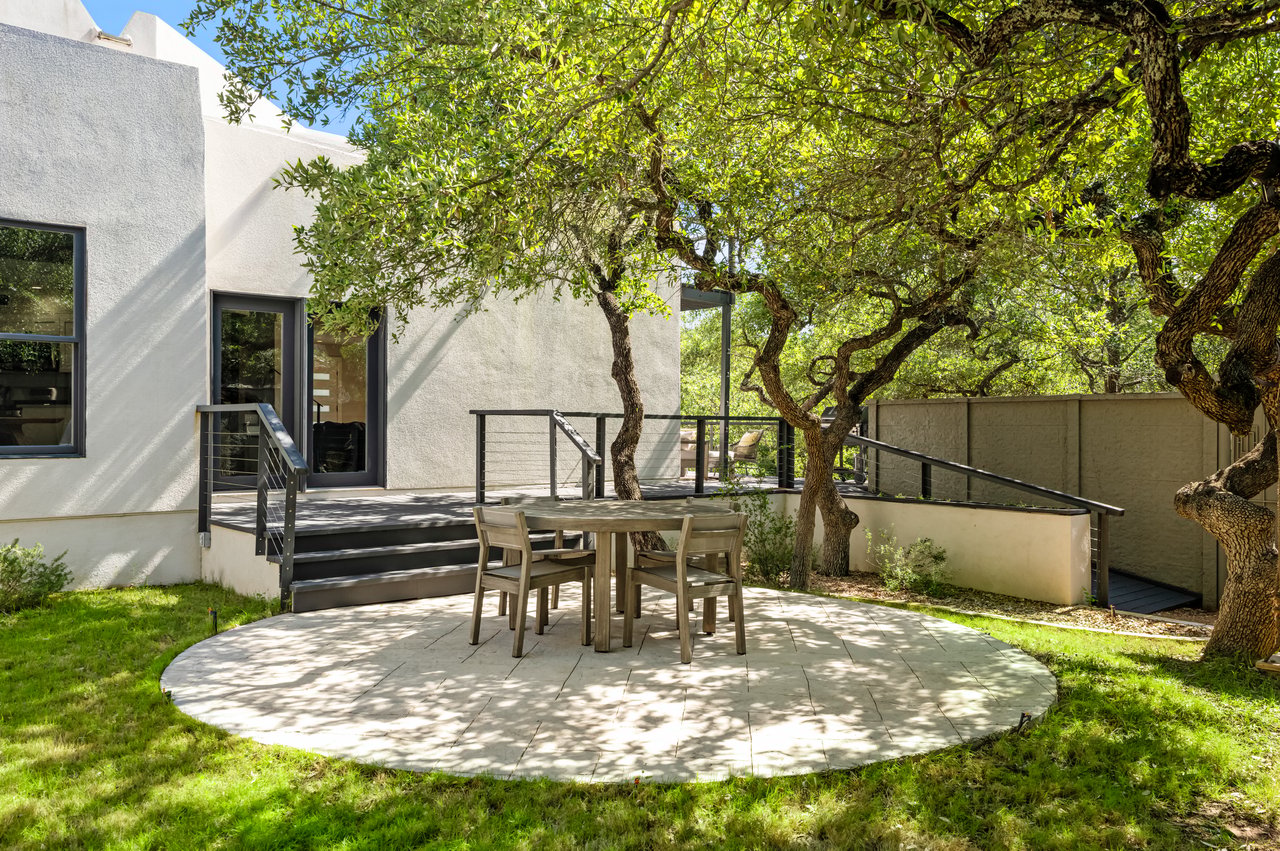 Stone-paved dining patio beneath leafy trees beside a modern white house and raised deck.