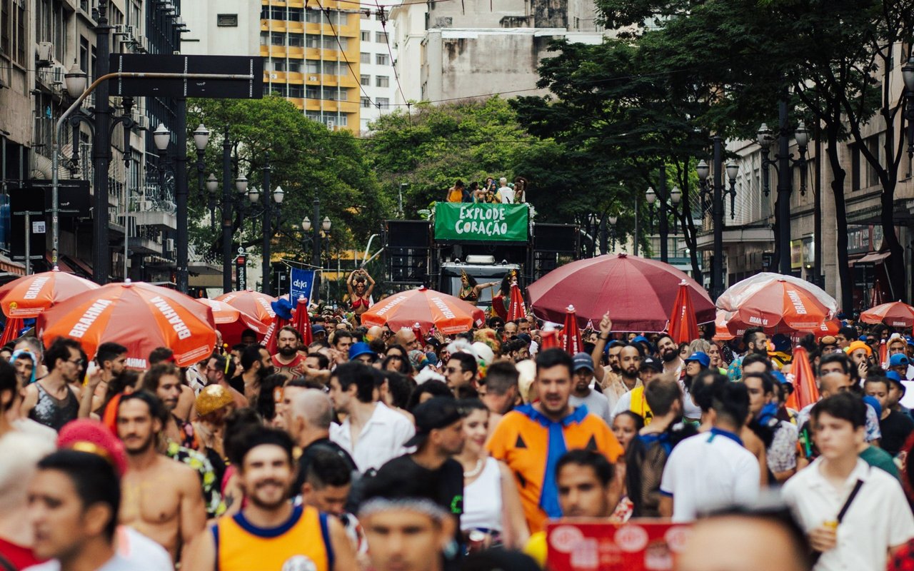 A crowded street festival beneath trees, with orange umbrellas and a green banner.