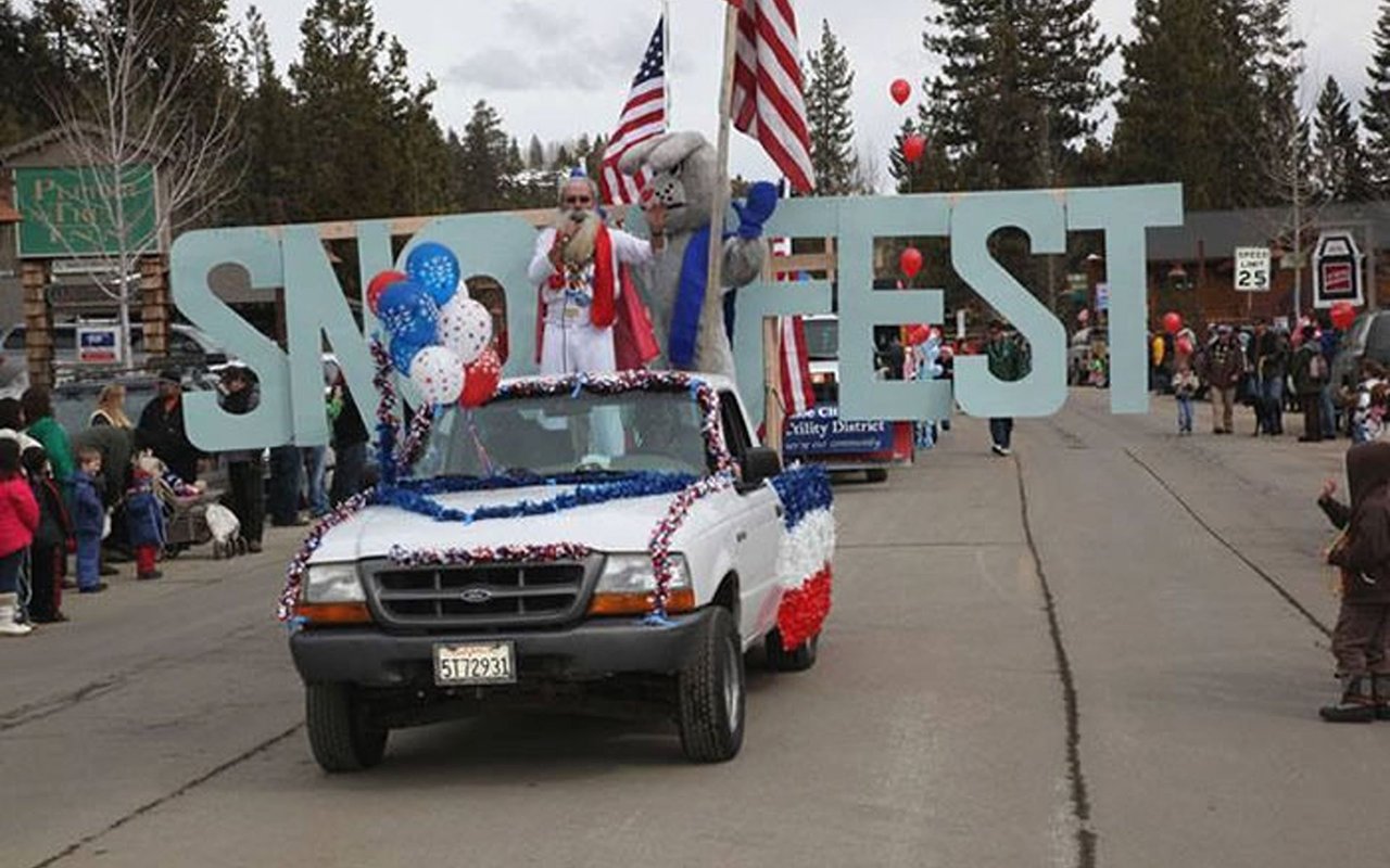 A patriotic parade pickup passes beneath large SNOWFEST letters as spectators line the street.