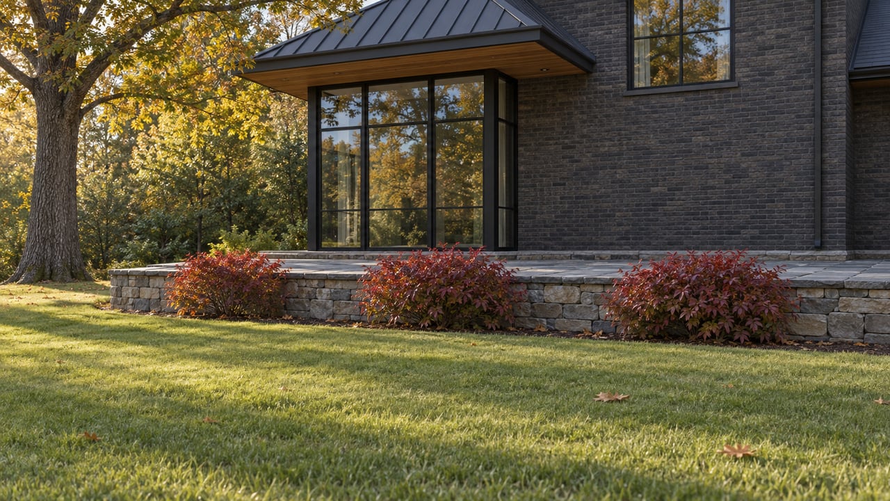 Charcoal-brick house corner above a stone terrace, with deep lawn and a gold-turning oak.