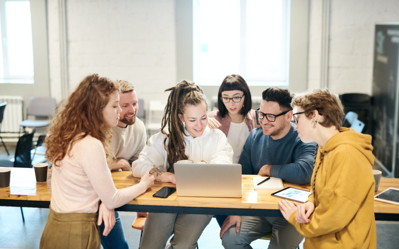 Six people collaborate around a laptop at a table.