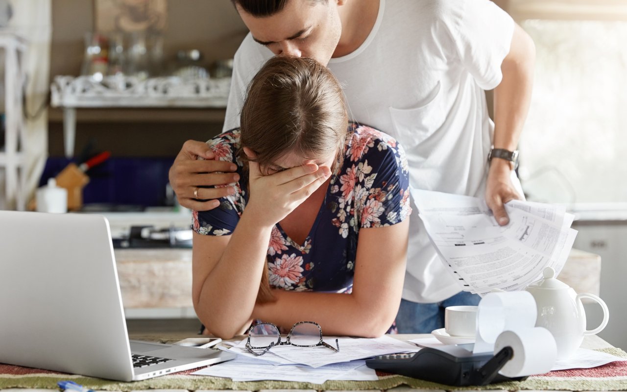 A man comforts a distressed woman seated at a kitchen table covered with paperwork.
