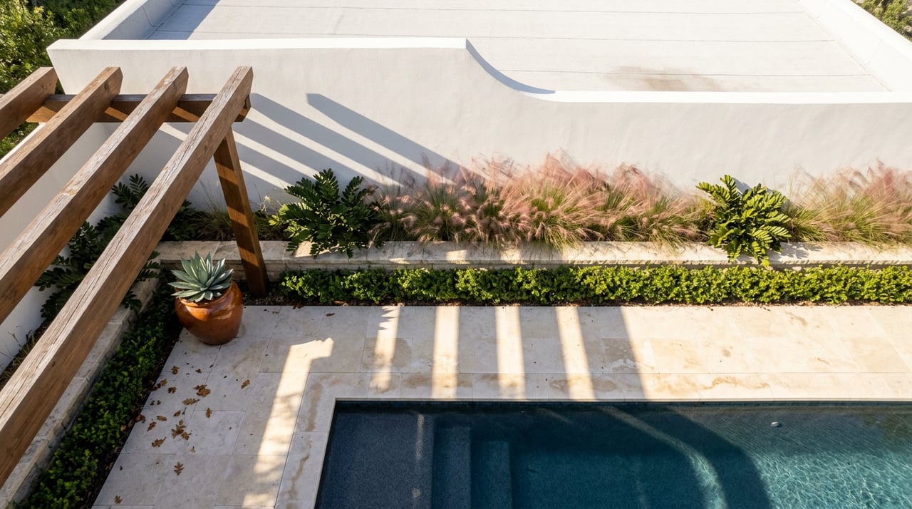 Overhead courtyard view of pergola shadows across pale stone paving beside a dark-tiled plunge pool, with an agave planter.