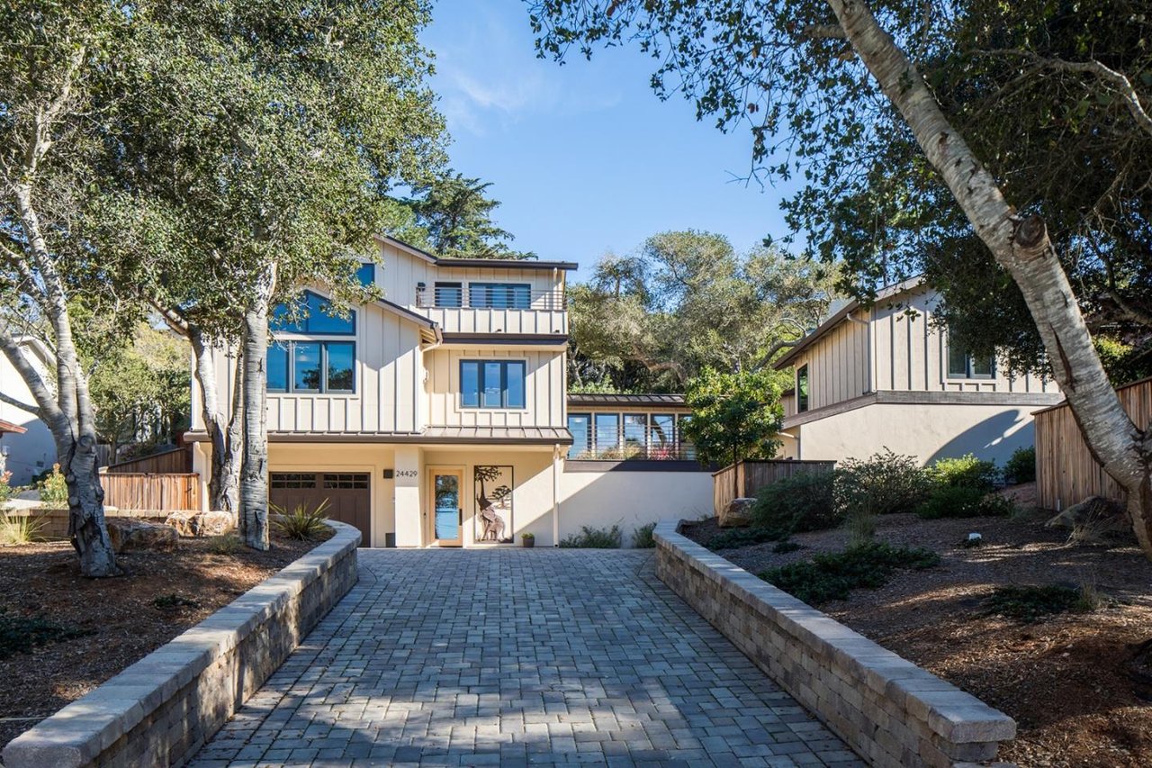 Modern two-story home with an upper balcony, reached by a paved driveway beneath mature trees.