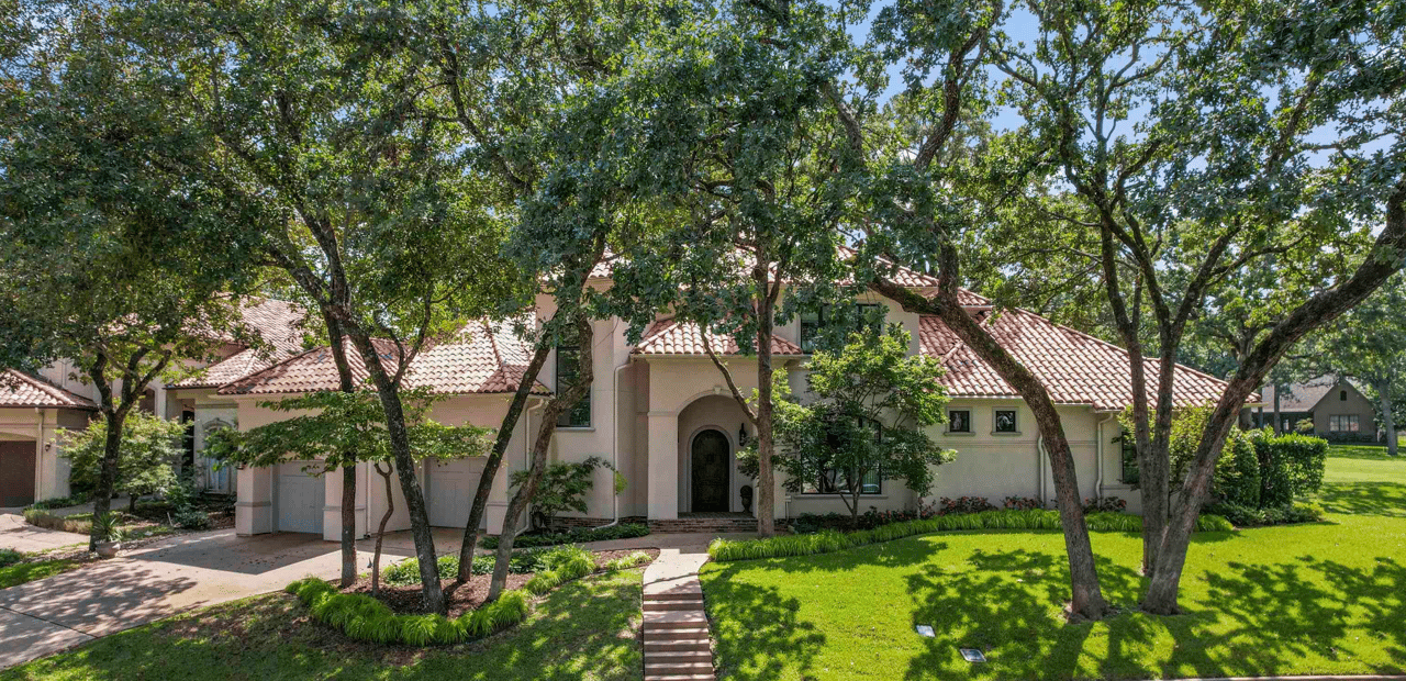 Front exterior of a two-story luxury home with terra cotta tile roof, arched entry, three-car garage, and manicured landscaping surrounded by mature oak trees in The Cascades neighborhood, Tyler, TX