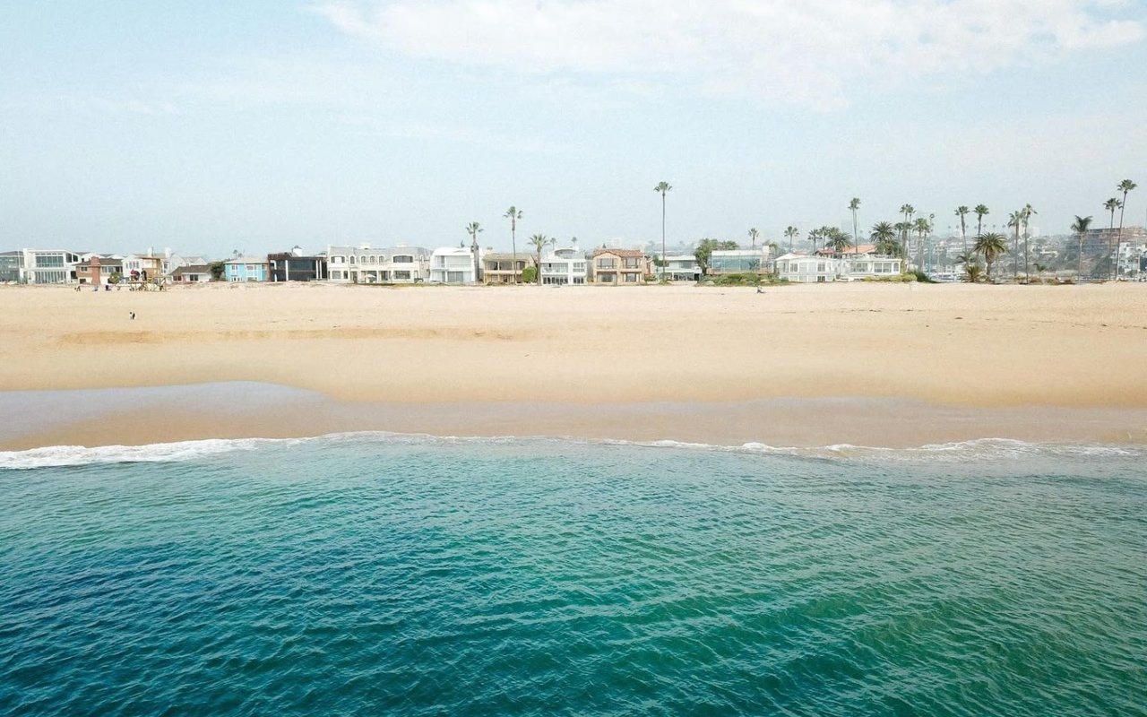 A coastal view showing turquoise ocean waves breaking on a wide, sandy beach lined with beachfront houses and palm trees.