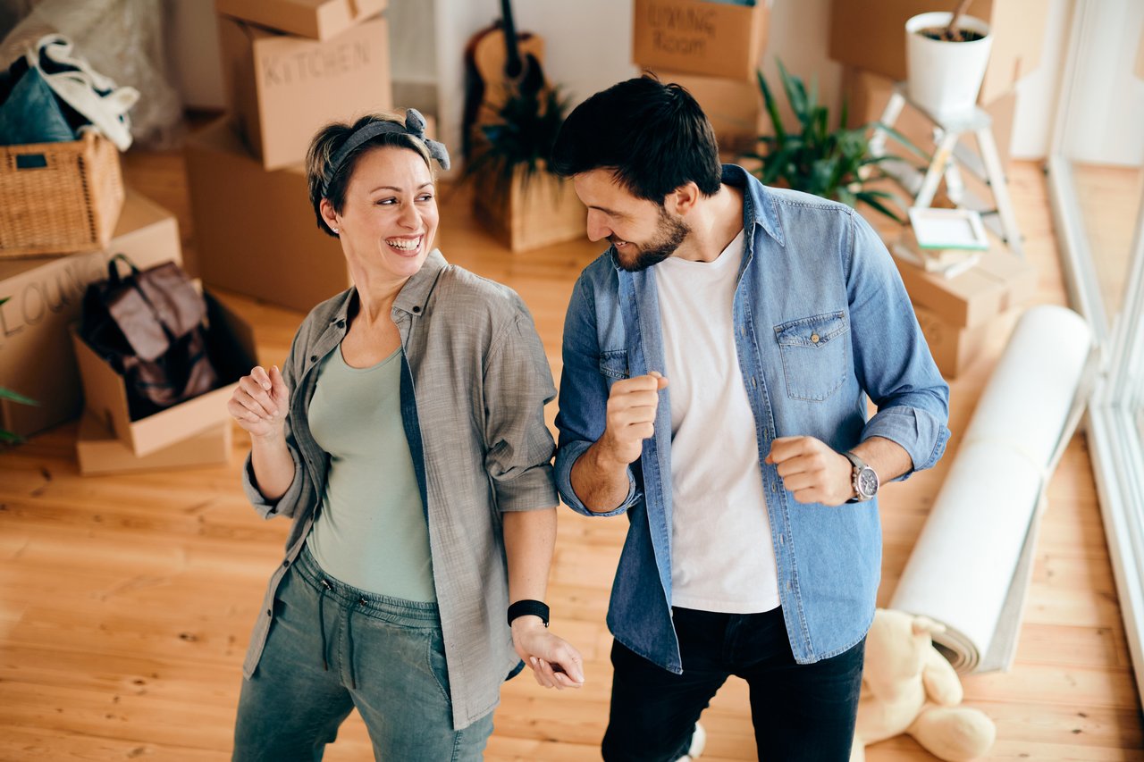 Happy couple dancing among moving boxes as they prepare to sell their home, celebrating a successful real estate transaction.