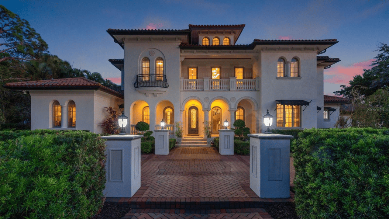 Two-story stucco home with terracotta roof, arched entry, and illuminated windows at dusk.
