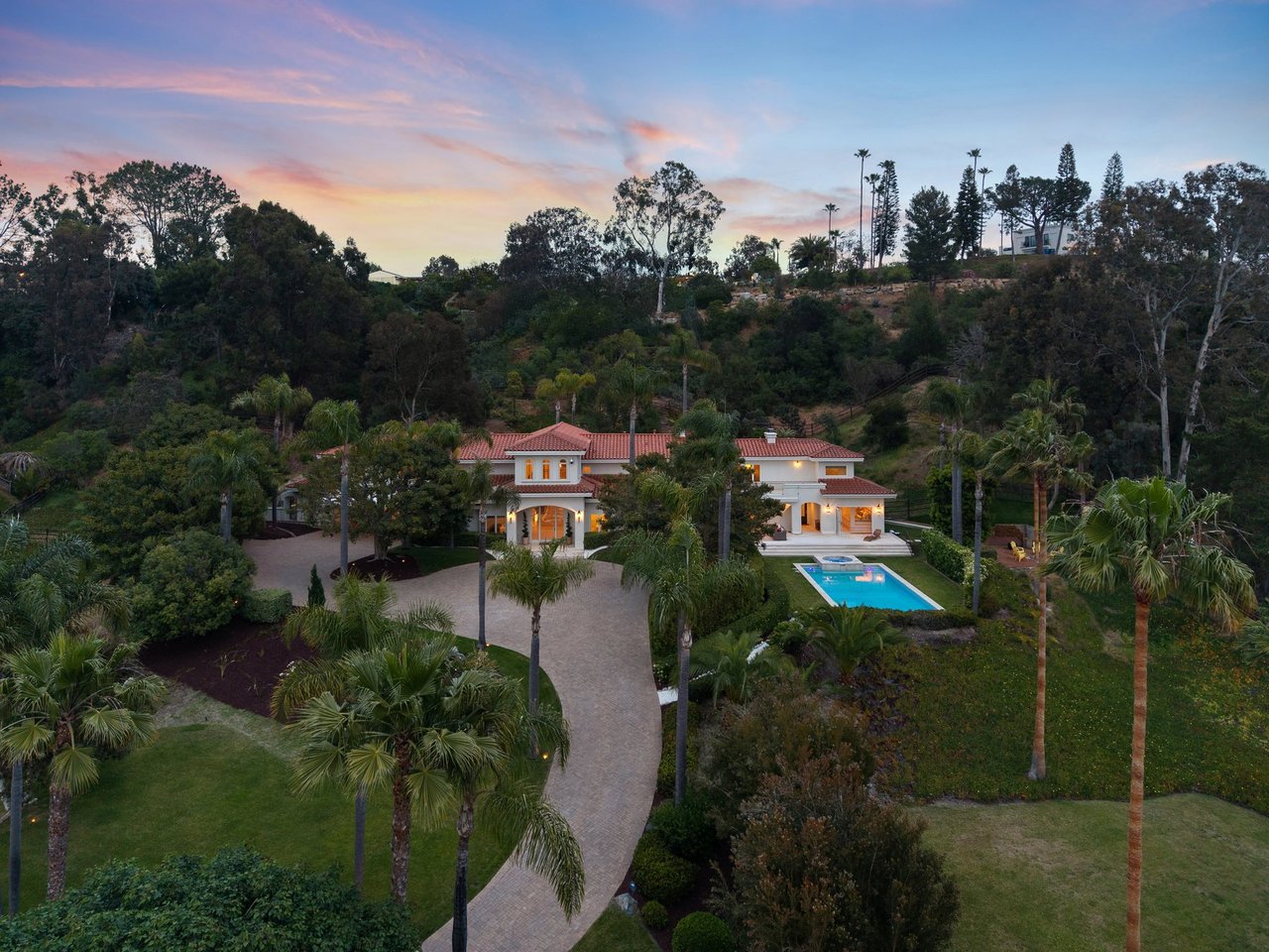 Aerial view of a white home with a pool, palm-lined grounds, and wooded hills at dusk.