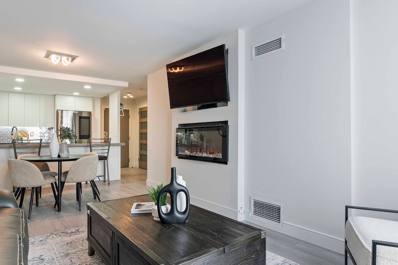 Open-plan condo living room with a wall-mounted TV, electric fireplace, and adjoining kitchen.