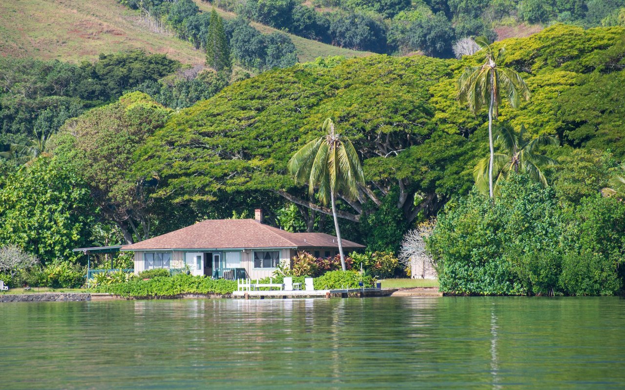 Waterfront home in Kokokahi beside calm water, framed by palm trees and a lush hillside.