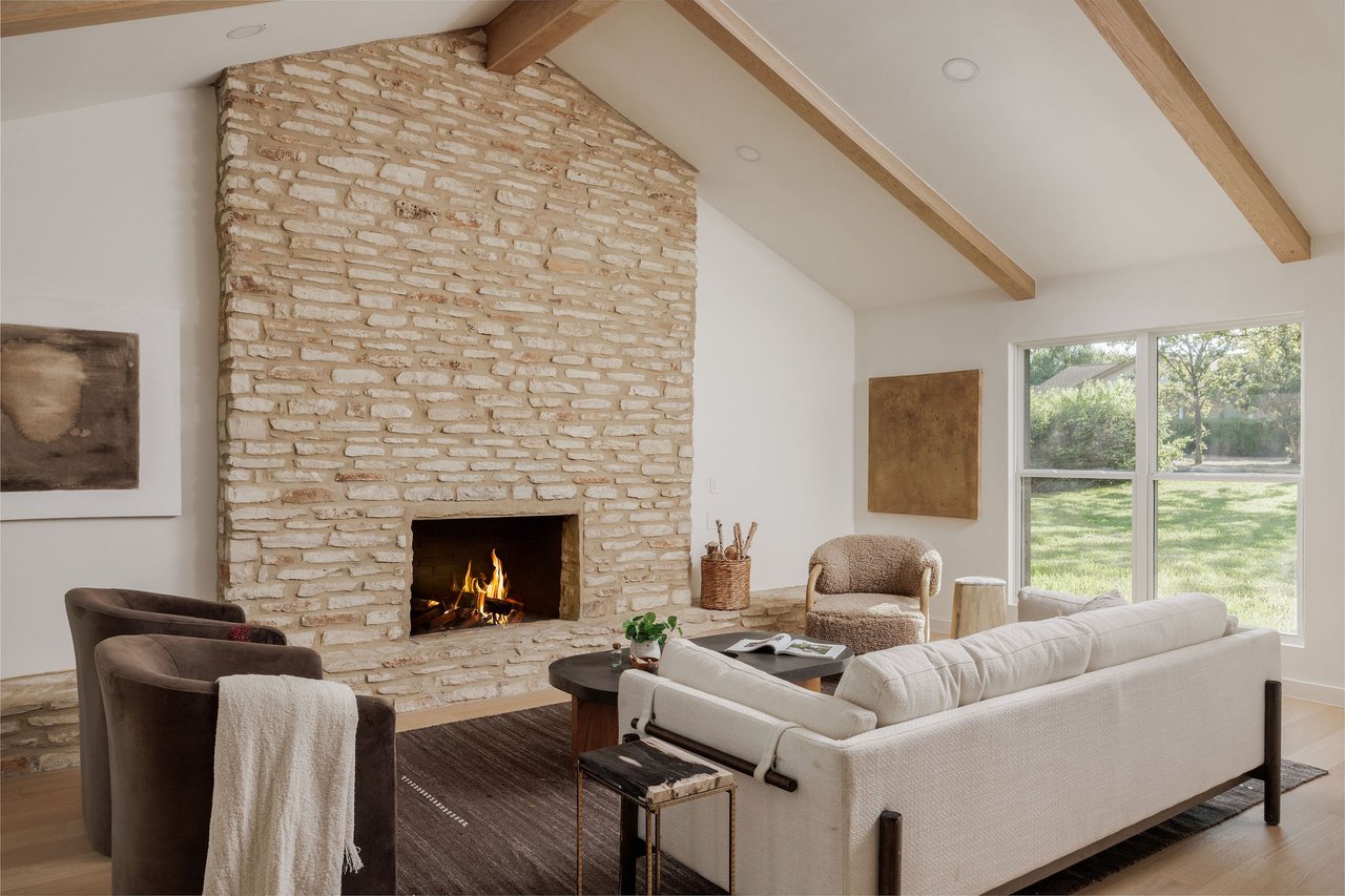 Living room with a tall limestone fireplace, exposed wood beams, and large windows overlooking a yard.