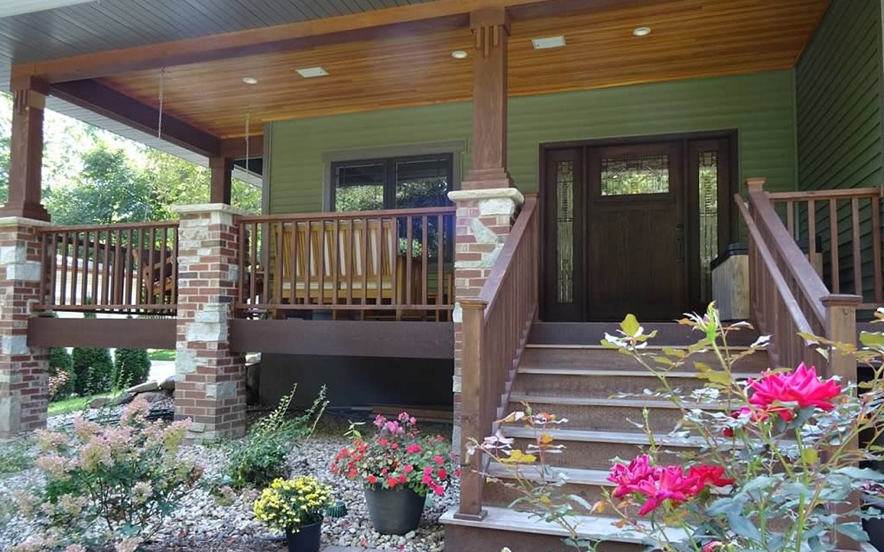 Covered front porch with wooden steps, brick columns, dark double doors, and colorful flower beds.