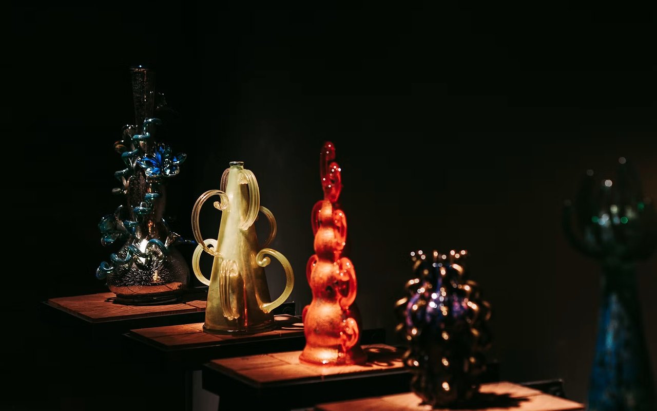 Colorful glass sculptures displayed on illuminated pedestals in a dark gallery.