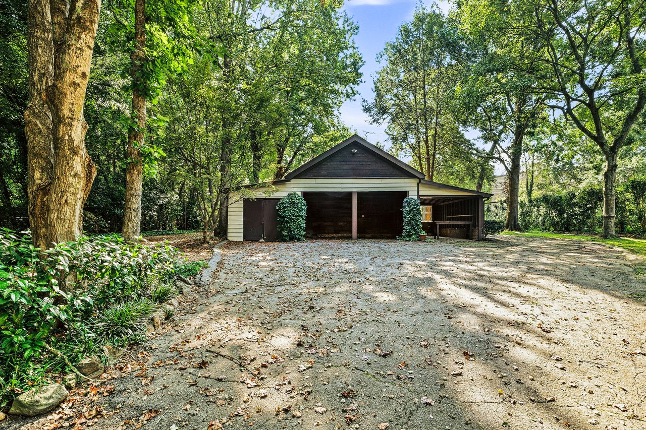 Detached garage with a covered carport behind a broad paved driveway and mature trees.