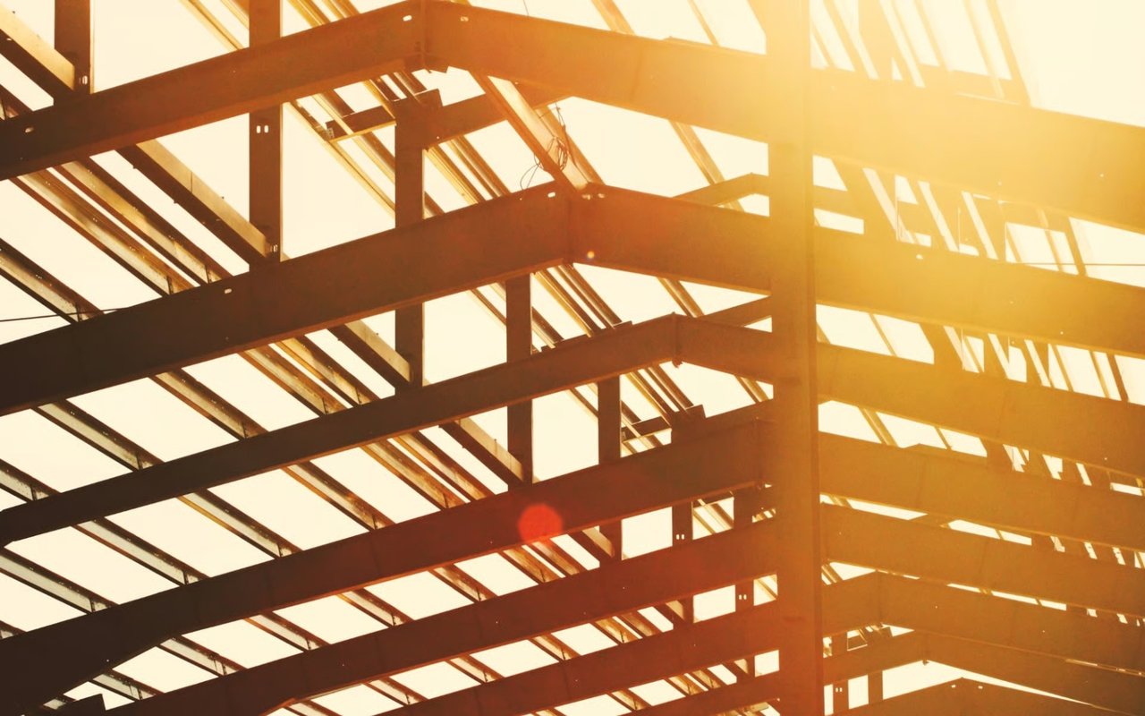 Industrial steel building framework silhouetted against a bright, warm golden hour sky with strong sunlight and a prominent lens flare.