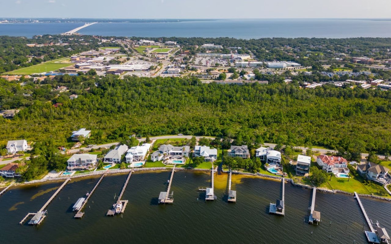 Aerial view of waterfront homes and docks, with a wooded area, town, and bay beyond.