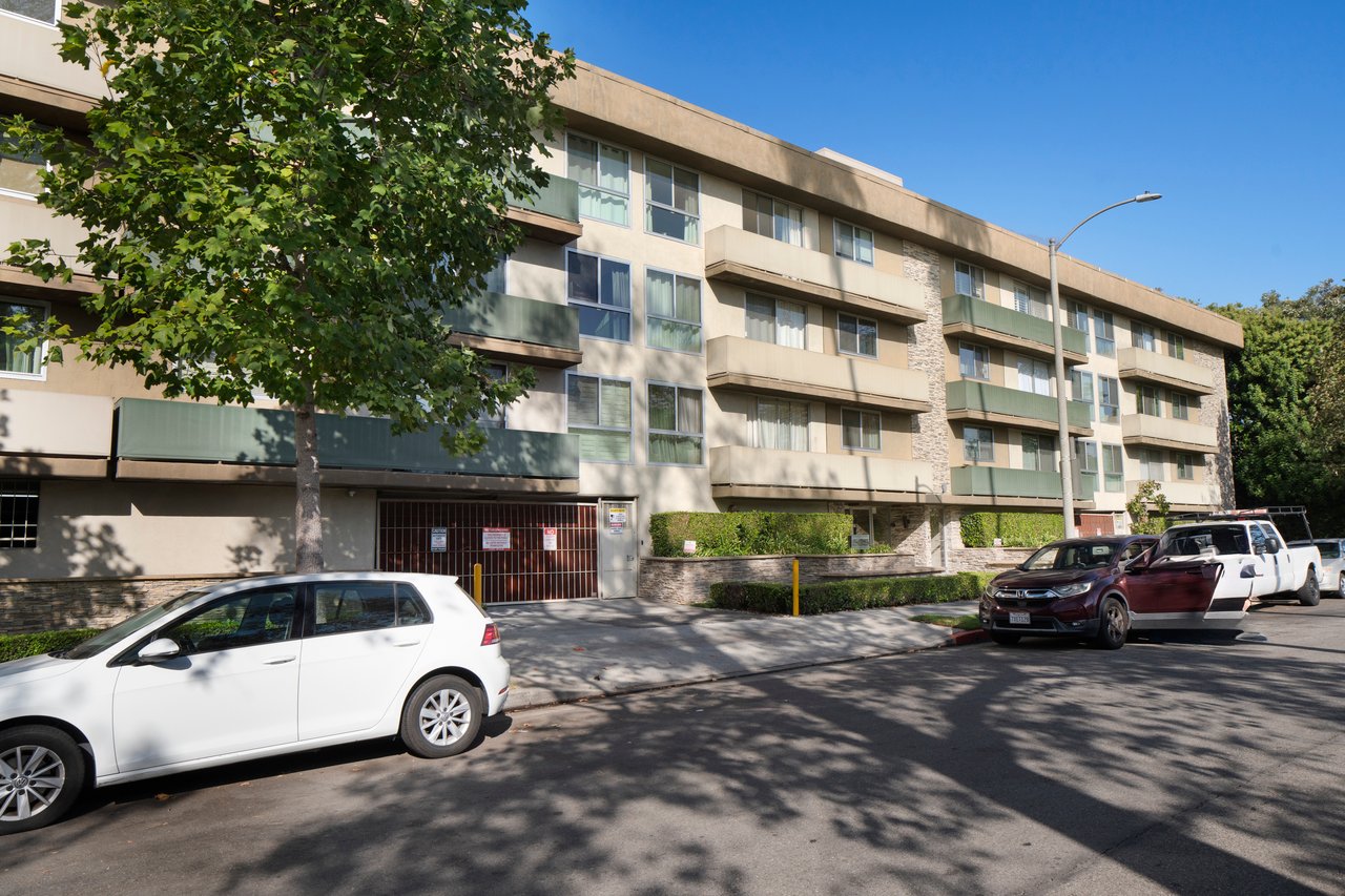 Multi-story apartment building with green balcony railings, partly screened by a leafy tree.