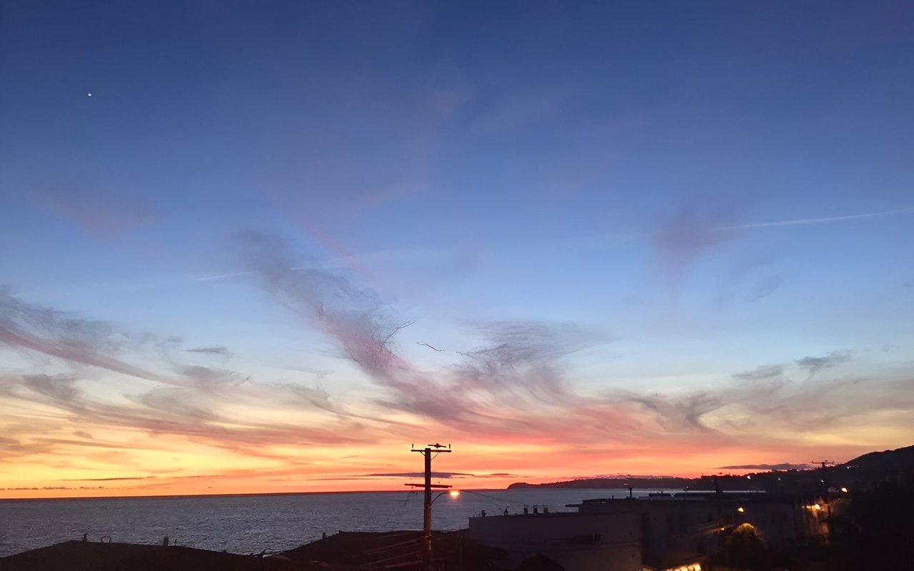 Winter sunset over the ocean, with pink clouds, coastal buildings, and a glowing horizon.