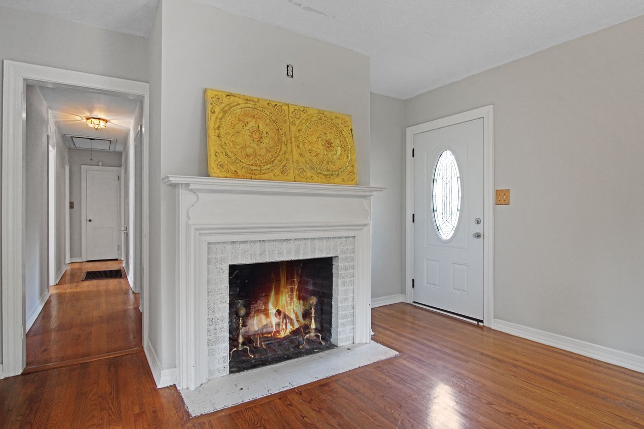 Bright hallway with wooden floors and a lit fireplace with a white mantel. Yellow artwork rests above. To the right, a door with an oval window. Cozy and inviting.