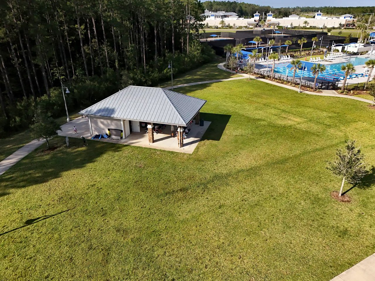 Aerial view of a community pool and pavilion beside a broad lawn and pine trees.
