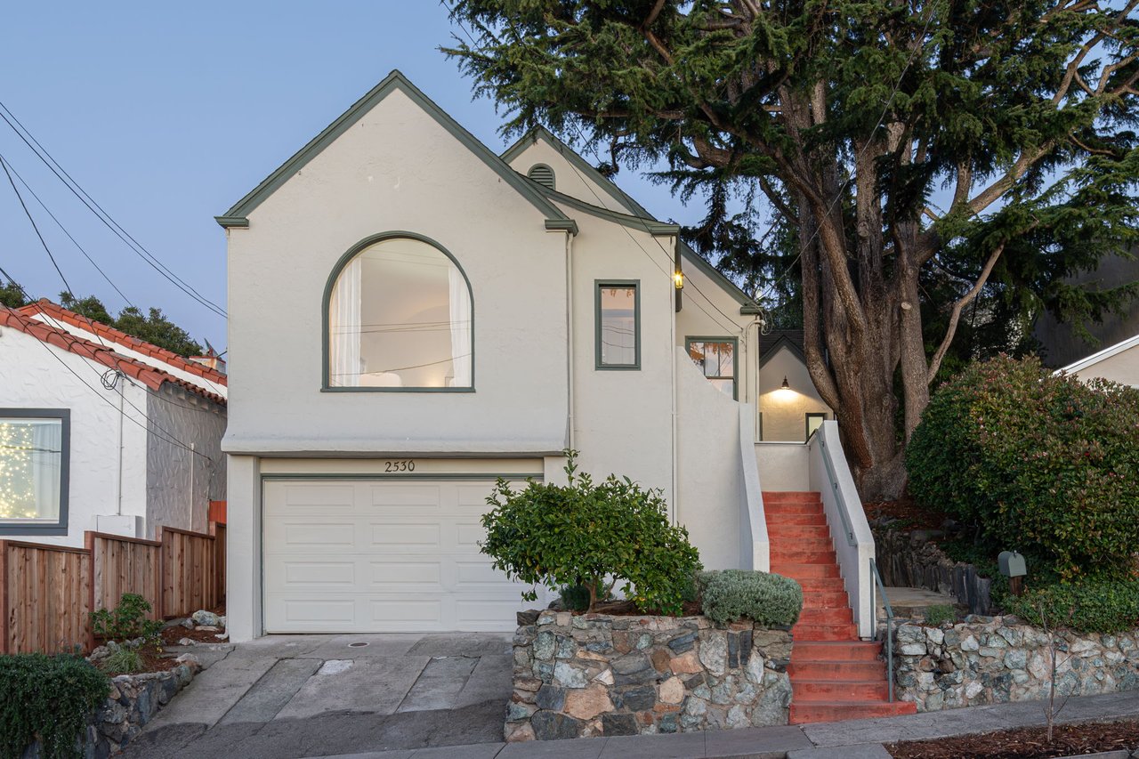 White two-story home with an arched window, attached garage, and red front steps beneath a large tree.