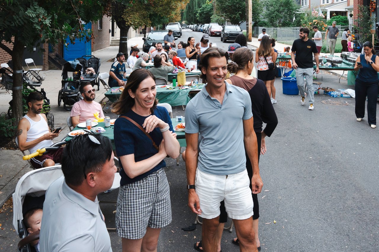 Neighbors mingle at a residential street block party beside tables of food and folding chairs.