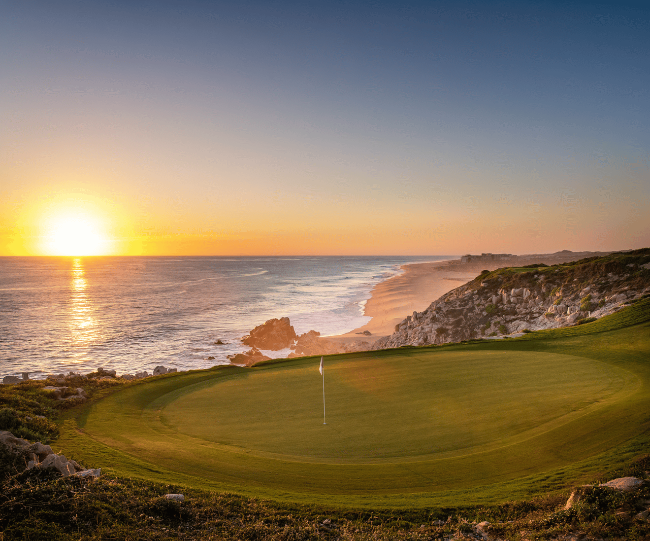 Coastal golf green overlooking a rocky beach and ocean at sunset.