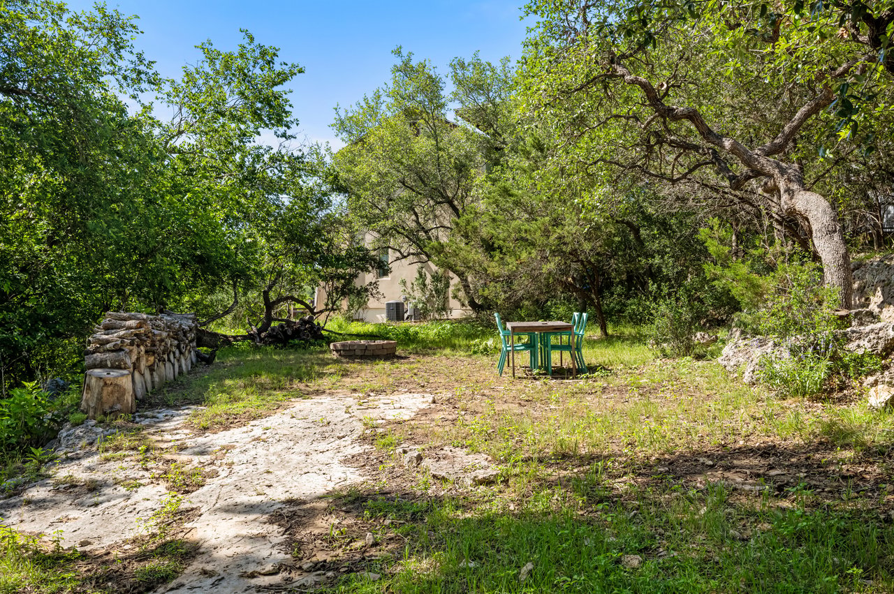 Wooded backyard with a stone fire pit, turquoise patio table and chairs, and stacked firewood.
